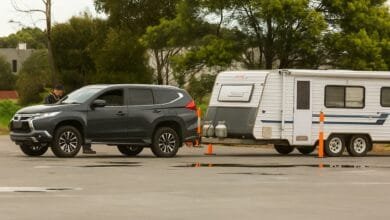 Un SUV gris tracte une caravane blanche, entourés d’arbres, cônes orange et surface mouillée.