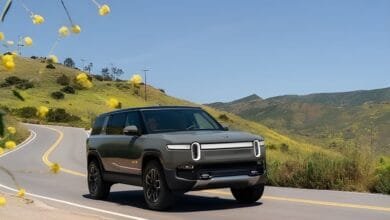 SUV électrique Rivian R1S gris sur route de montagne avec fleurs jaunes et collines verdoyantes.RéessayerClaude peut faire des erreurs. Assurez-vous de vérifier ses réponses.