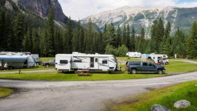 Une voiture tracte une grande caravane dans un camping alpin entouré de sapins et montagnes majestueuses.