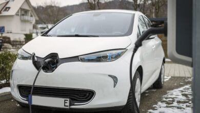 Voiture électrique blanche Renault Zoé en charge à domicile, câble de recharge branché, stationnée dans une allée résidentielle enneigée avec maisons en arrière-plan.Réessayer
