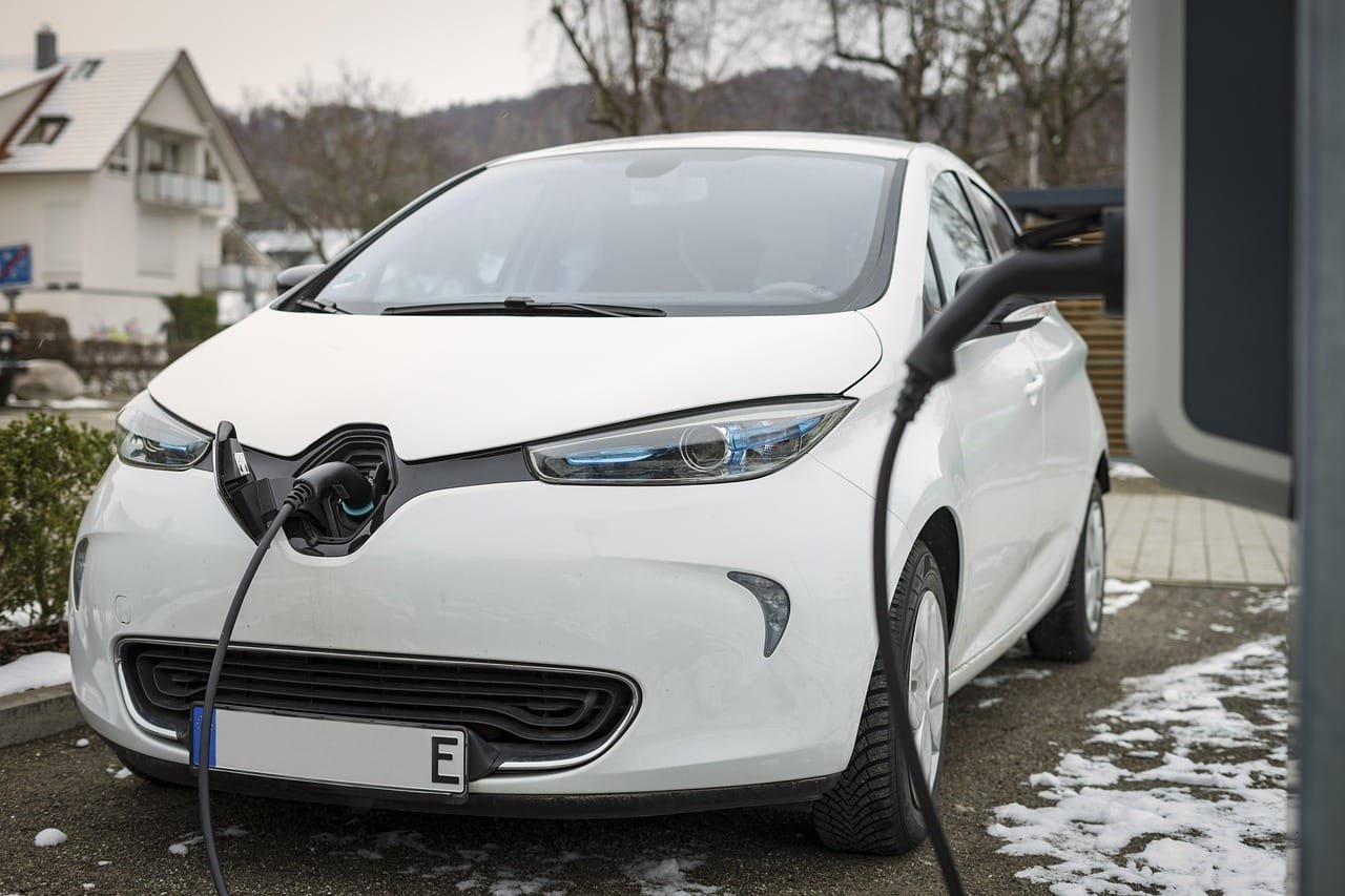 Voiture électrique blanche Renault Zoé en charge à domicile, câble de recharge branché, stationnée dans une allée résidentielle enneigée avec maisons en arrière-plan.Réessayer
